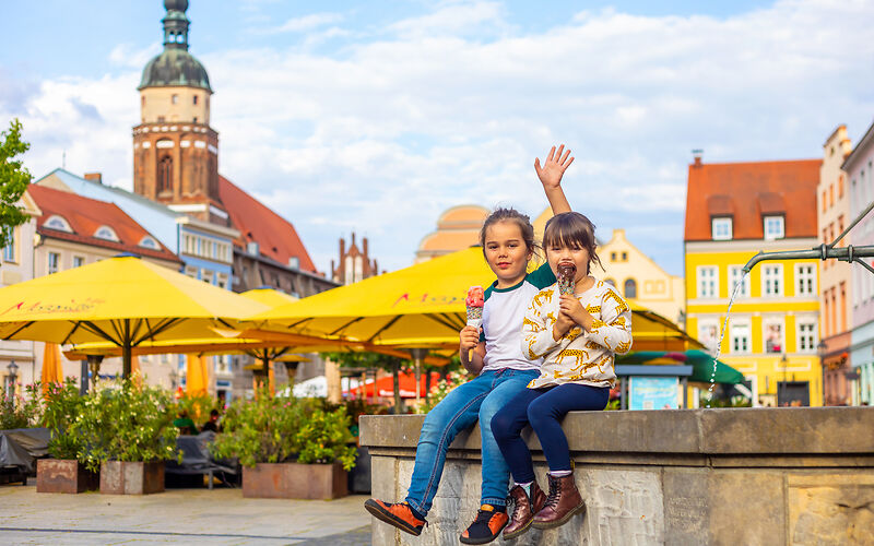 Auf dem Foto ist der Altmarkt der Stadt Cottbus zu sehen. Zwei kleine Mädchen sitzen auf dem historischen Brunnen und essen ein Eis. Im Hintergrund befindet sich der Turm von der Oberkirche.