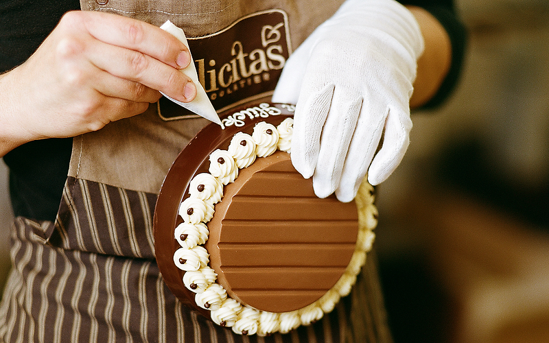 Auf dem Foto ist eine Torte zu sehen, die von einer Frau liebevoll verziert wird. Das Logo „Felicitas“ ist gut sichtbar auf ihrer Schürze. 