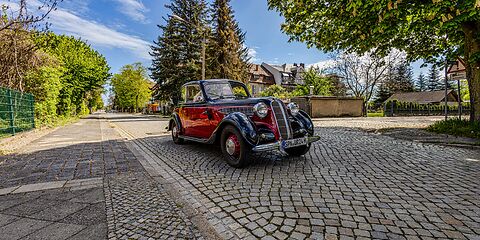 Ein schwarz-roter Oldtimer fährt über die Kopfsteinpflasterstraße durch Forst (Lausitz). Die malerische, historische Straße ist von charmanten Gebäuden gesäumt, die dem Bild eine nostalgische Atmosphäre verleihen, während der Oldtimer gemächlich die Szenerie durchquert.