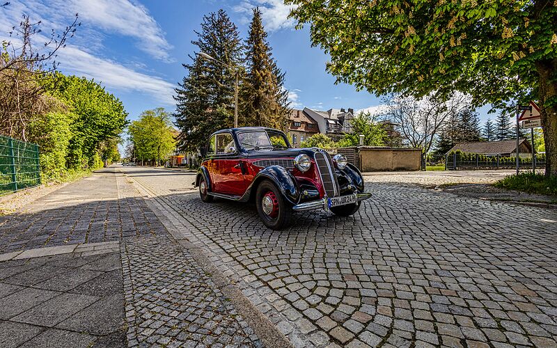 Ein schwarz-roter Oldtimer fährt über die Kopfsteinpflasterstraße durch Forst (Lausitz). Die malerische, historische Straße ist von charmanten Gebäuden gesäumt, die dem Bild eine nostalgische Atmosphäre verleihen, während der Oldtimer gemächlich die Szenerie durchquert.