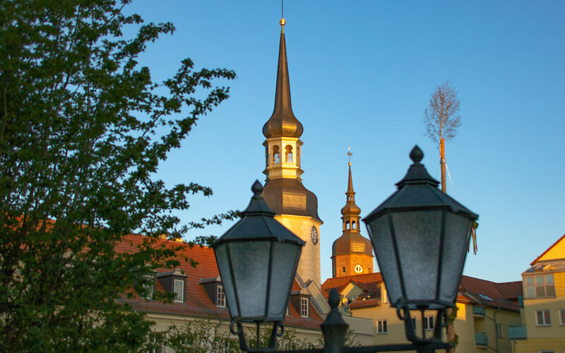 Ein malerischer Blick auf die Türme der Stadt Spremberg am frühen Morgen, sanft von ersten Sonnenstrahlen erleuchtet. Im Vordergrund steht eine alte Laterne, die dem Bild einen charmanten, nostalgischen Touch verleiht.