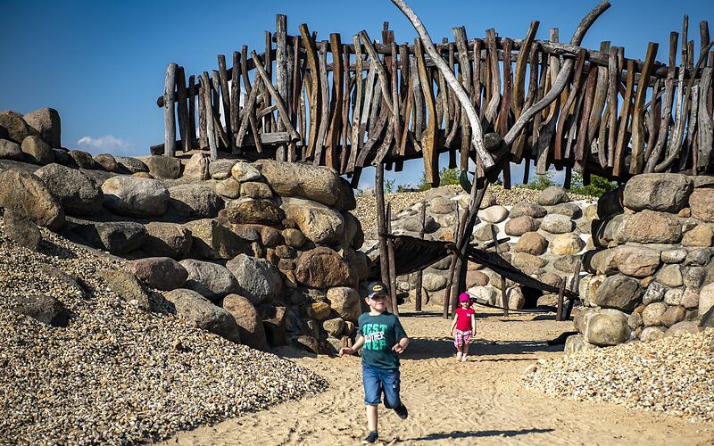 Auf dem Foto sind zwei Kinder auf einem Spielplatz mit einer Steinmauer und einer Holzbrücke zu sehen. 