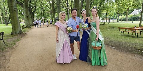 Auf einem Weg im Rosenkarten, an welchem links und rechts Bäume stehen, steht die Forster Rosenkönigin zusammen mit einem Roporter vom rbb, der Peitzer Teichnixe und der Spremberger Spreenixe.