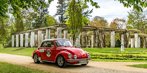 Ein roter Oldtimer-Volkswagen parkt malerisch im Ostdeutschen Rosengarten in Forst. Im Hintergrund ist der historische Pergolenhof zu sehen, der die Szene mit seiner eleganten Architektur prägt.