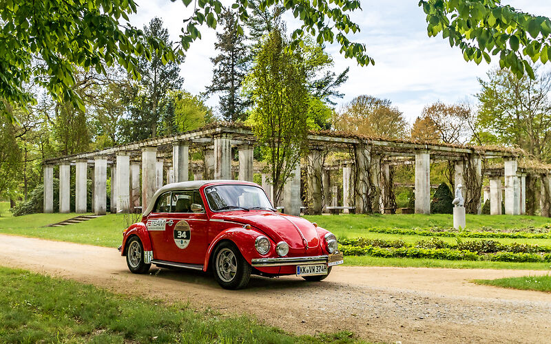 Ein roter Oldtimer-Volkswagen parkt malerisch im Ostdeutschen Rosengarten in Forst. Im Hintergrund ist der historische Pergolenhof zu sehen, der die Szene mit seiner eleganten Architektur prägt.